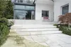 A modern house with a patio and stairs leading up to the entrance. The patio and stairs are made of light-colored stone tiles. There is greenery and a small tree around the patio. Anteprima dell’immagine prodotto