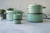 A collection of green cast iron pots and pans are arranged on a textured gray surface, with some greenery in the background. Produktbild-Vorschau