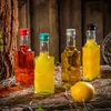 Four small glass bottles filled with colorful liquids, a lemon, and straw-like material on a wooden surface. Produktbild-Vorschau
