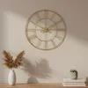A gold-colored wall clock with Roman numerals is displayed on a light beige wall above a wooden surface. A white vase with dried reeds and a small potted plant on a stack of books are also visible. Voir les images du produit