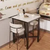 A small bar table with two stools in a cafe or dining area, with a display case of desserts in the background. Náhľad obrázku produktu