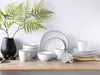 A collection of white ceramic dishes and mugs with black trim, arranged on a wooden shelf with a green fern in a black vase. Náhľad obrázku produktu