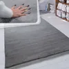 A hand pressing into a soft, gray, high-pile rug, showcasing its plushness and texture. The rug is laid out on a light-colored wooden floor, with a bookshelf and part of a living room visible in the background. Produktbild-Vorschau