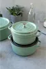 A stack of green cast iron pots on a gray surface, with a plant and a bottle in the background. Produktbild-Vorschau