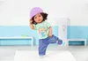 A doll is standing on a rug in a room with blue and white walls and white benches. Produktbild-Vorschau