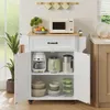 A white kitchen cart with a wooden countertop is shown in a well-lit room. The cart has two doors open, revealing shelves with kitchen appliances and cookware. A plant is visible to the left, and a window with frosted glass is behind it. Podgląd zdjęcia produktu