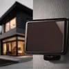 A solar-powered LED spotlight is mounted on a textured wall, with a modern house blurred in the background. Produktbild-Vorschau