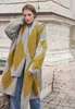 A woman wearing a knitted cardigan and jeans, standing outdoors near a stone structure. Produktbild-Vorschau