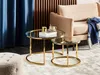 A living room scene featuring two round glass and gold coffee tables, a beige armchair with a blue and white striped throw blanket, and a plush ottoman. Náhľad obrázku produktu