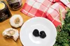 A close-up of two black truffles on a white plate, with garlic, jars of truffle products, and greens in the background. Podgląd zdjęcia produktu
