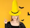 A woman wearing a large yellow witch hat and a black dress, with black paper bats on an orange background. Produktbild-Vorschau