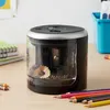 A black electric pencil sharpener with a clear container sits on a wooden desk, surrounded by colored pencils and notebooks. A blue bin is visible in the background. Anteprima dell’immagine prodotto