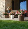 Several potted plants with pink and red flowers are arranged on a patio in front of a stone building and a wooden bench. Náhled obrázku produktu