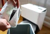 A person is holding a bag of dust and emptying it into a bin next to a white automated cat litter box. Produktbild-Vorschau