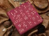 A close-up of a red advent calendar with gold numbers and festive designs, set against a textured brown burlap background with natural elements like stones and wood. Anteprima dell’immagine prodotto