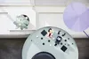 Two LEGO BrickHeadz figures, Cruella and Maleficent, are arranged on a round, light blue table with scattered black and white square LEGO pieces. A succulent plant in a pot sits on a white shelf in the background, and a large, pale purple disc is visible t Anteprima dell’immagine prodotto
