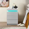 A white nightstand with three drawers and LED lighting is placed next to a bed. Produktbild-Vorschau