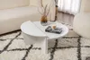 A white coffee table is placed on a rug in a living room setting, with a sofa and curtains in the background. Produktbild-Vorschau