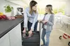 A mother and daughter are in a kitchen, with the mother disposing of trash. Produktbild-Vorschau