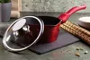 A red saucepan with a glass lid is placed on a dark slate coaster, with a bamboo mat and a bowl of rice in the background. Anteprima dell’immagine prodotto