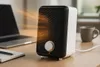 A small black and white space heater is shown on a wooden desk next to a laptop and a notebook. Produktbild-Vorschau