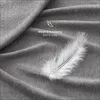 A close-up shot of a soft, gray fabric with a white feather resting on it. Text overlays indicate the product is soft and cozy. Produktbild-Vorschau