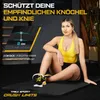A woman is sitting on a yoga mat in a gym-like setting, with text overlays highlighting product features. Produktbild-Vorschau