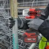 A person wearing work gloves is using a red and black cordless impact wrench on a metal pole, with scaffolding in the background. Podgląd zdjęcia produktu