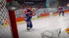A hockey player in a blue and orange uniform is skating on an ice rink, about to hit a puck that is flying towards the camera. The background shows a stadium filled with spectators. Produktbild-Vorschau