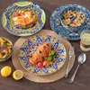 A table setting with colorful, bohemian-style ceramic plates and bowls filled with food, alongside cutlery and a glass of water. Produktbild-Vorschau