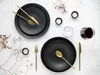 A flat lay of a dinner setting with black plates, gold cutlery, and wine glasses. Produktbild-Vorschau