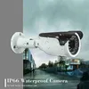 A security camera mounted on a wall with a background showing a street scene with modern houses and people walking. Náhľad obrázku produktu