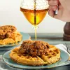 A hand is pouring honey from a honeycomb-shaped dispenser onto chicken and waffles. Produktbild-Vorschau