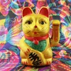 A golden Maneki Neko cat figurine sits in front of a colorful, abstract background. Produktbild-Vorschau