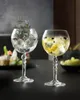 Two crystal gin tonic glasses filled with ice and garnishes, placed on a dark table with a blurred background. Produktbild-Vorschau