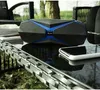 A Bluetooth speaker and a smartphone are placed on a glass table outdoors, with chairs and foliage in the background. Produktbild-Vorschau