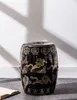 A black ceramic garden stool with gold dragon motifs sits on a gray rug in front of a white wall with shadows. Anteprima dell’immagine prodotto