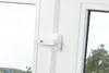 A close-up view of a white window handle with a small white device attached to it, showing a few vertical lines and a small circular icon. The background shows a slightly out-of-focus view of trees and sky through the window. Produktbild-Vorschau