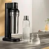 A soda maker with a stainless steel bottle, a glass bottle of water, and a glass of water on a countertop. Produktbild-Vorschau