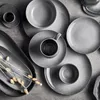 A top-down view of a set of grey ceramic plates and cups arranged on a grey textured surface. Anteprima dell’immagine prodotto
