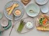 A collection of ceramic plates and bowls filled with various snacks and dips, arranged on a textured surface. Produktbild-Vorschau