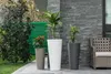 Three planters with plants are arranged on a patio next to a stone wall and a chair. Náhľad obrázku produktu