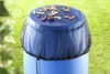 A blue rain barrel covered with a black mesh net, with leaves on top of the net, set in a grassy outdoor environment. Náhľad obrázku produktu