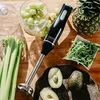 A black immersion blender is placed on a wooden surface surrounded by ingredients for making a smoothie, including grapes, celery, avocado, pineapple, and arugula. Produktbild-Vorschau