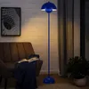 A blue floor lamp is shown in a living room setting next to a chair and a plant. Produktbild-Vorschau