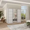 A bedroom scene featuring a large white sliding door wardrobe with mirrored panels, reflecting a floral wallpaper and a bed. The room has wooden flooring and is decorated with plants and a chair. Produktbild-Vorschau