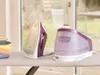 A Braun steam generator iron and its water tank are placed on an ironing board next to a stack of folded clothes. The background shows a blurred view of a room with a window and outdoor scenery. Produktbild-Vorschau