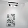 A black ceiling light fixture with two adjustable spotlights is mounted on a white ceiling. Below the light fixture, a framed piece of abstract art with circular patterns is hanging on a white wall. Produktbild-Vorschau