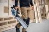 A person is holding a circular saw in a workshop setting with wood and construction materials in the background. Produktbild-Vorschau
