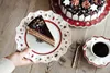 A slice of chocolate cake on a festive plate, with a cup of coffee and a whole cake in the background. Náhled obrázku produktu